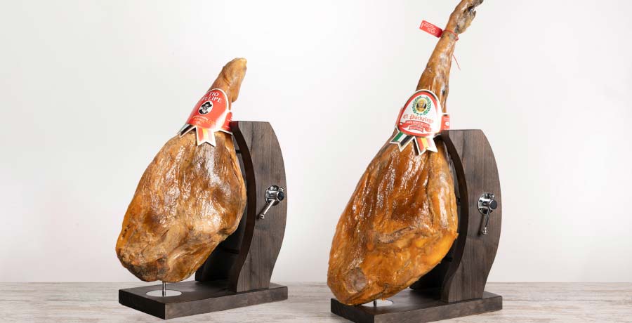 Jamón serrano y jamón ibérico colocados en soportes jamoneros, resaltando sus diferencias en tamaño, textura y etiquetado.